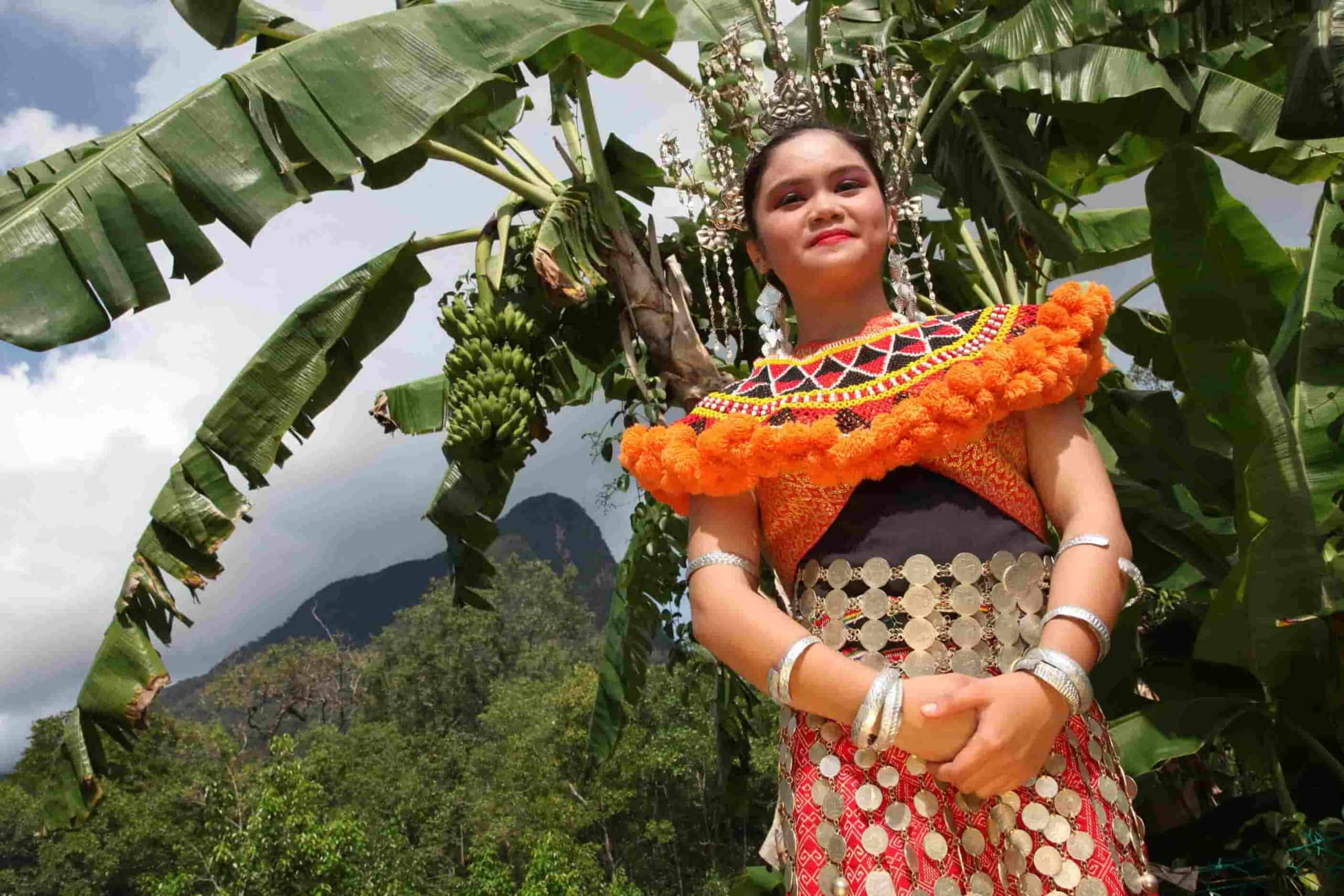 seorang gadis orang asli Sarawak dalam pakaian tradisi budaya berdiri di hadapan pokok pisang. Di latar belakang adalah bukit dan pokok.