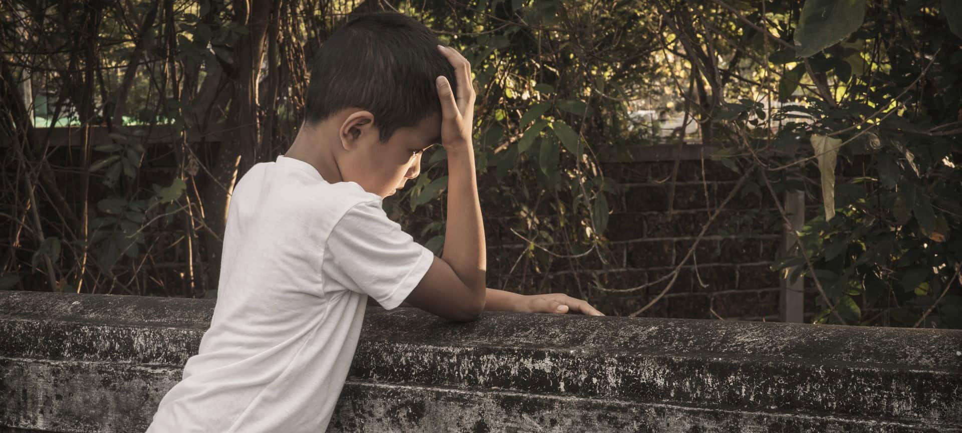 A boy stands against the wall with his head in his palm. The boy looks sad.