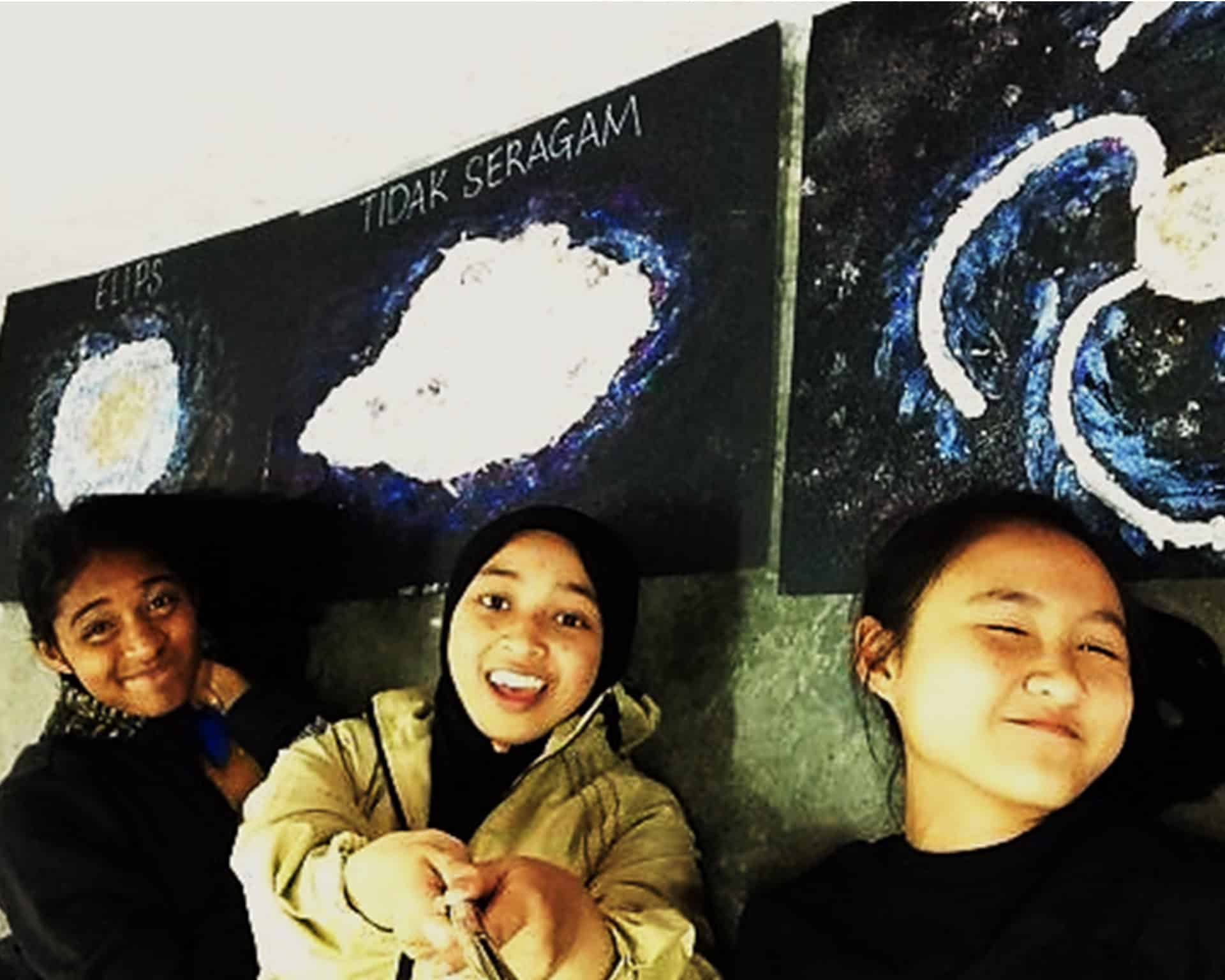 Three girls stand in front of a black board.