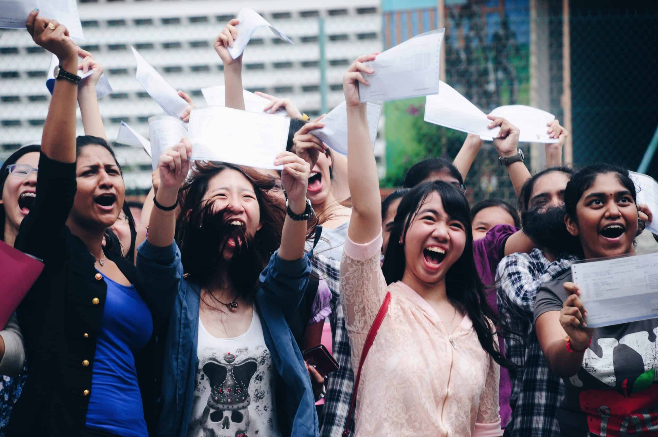 a group of girls hold up their exam results. The girls are smiling and laughing.