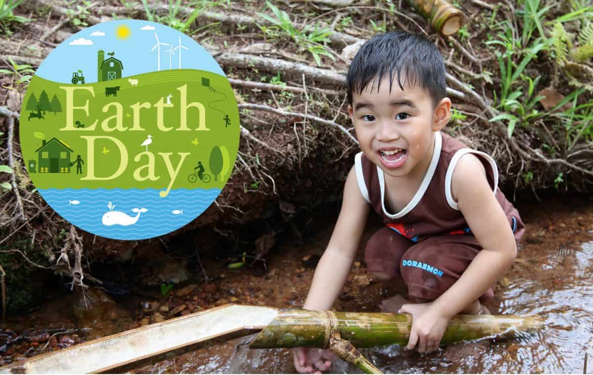 a young boy holds a bamboo stick in a river. Next to him in a the Earth Day logo.