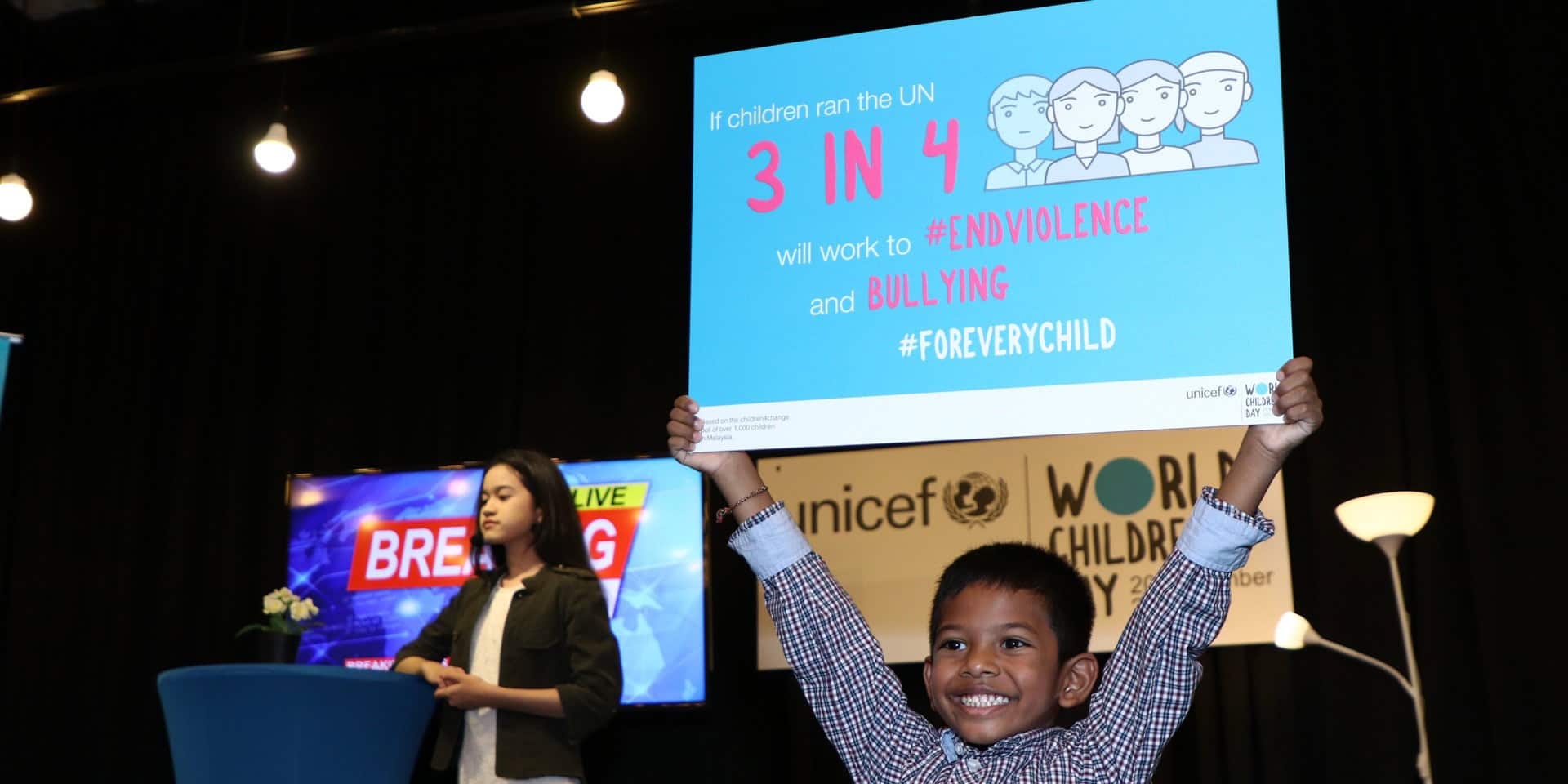 A boy holds up a placard that says If children ran the UN, 3 in 4 will work to end violence and bullying for every child. Behind him a girl presenter stands infront of a TV screen with the message Breaking News.