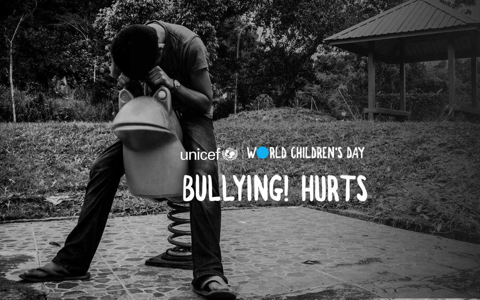 A dejected looking boy sits in the playground with his head on his hand. On the photo is the phrase Bullying hurts!