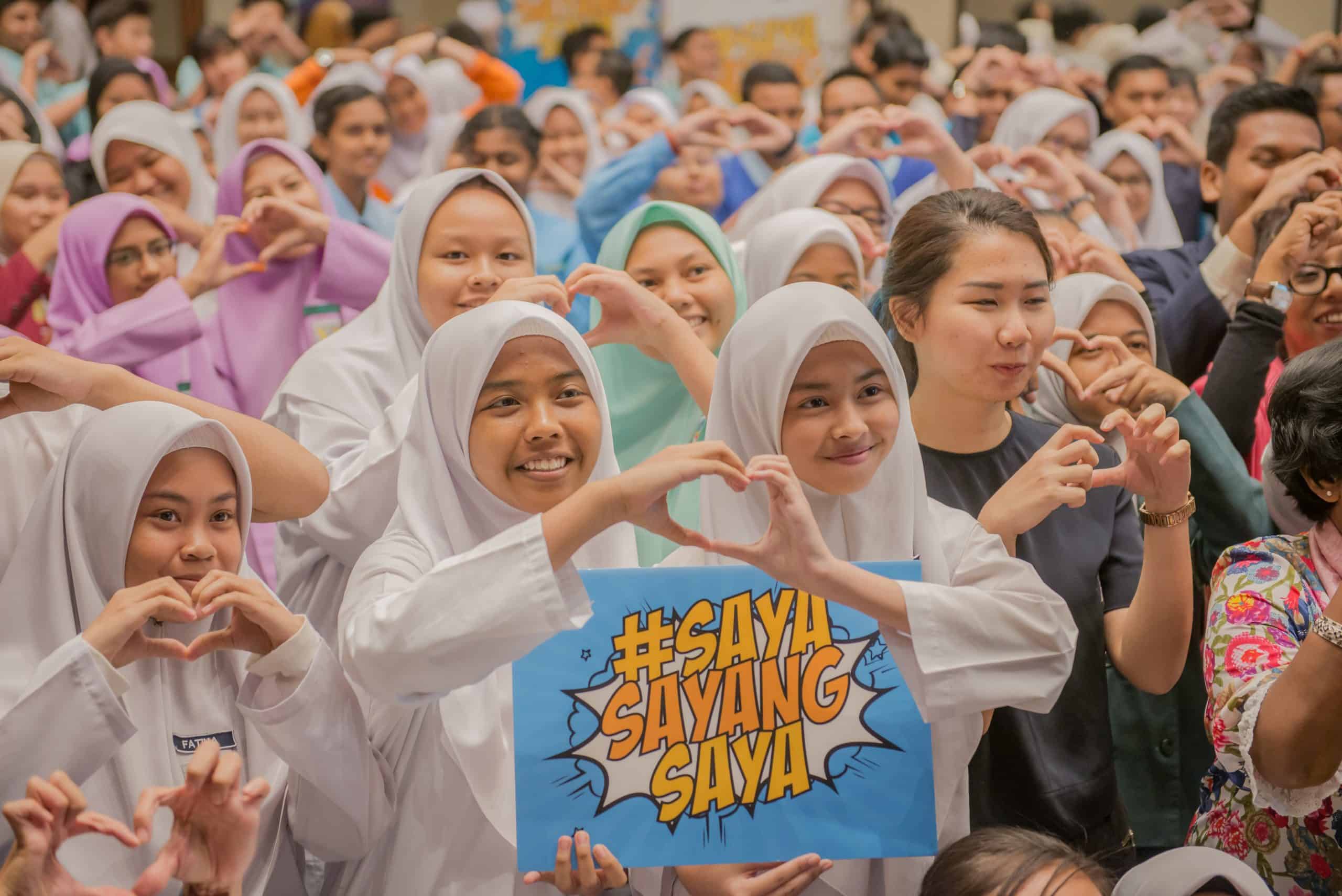 a group of school students showing the heart sign with their two hands as a symbol calling for sex education to protect them.. Two girls hold a sign board with the words Saya Sayang Saya.