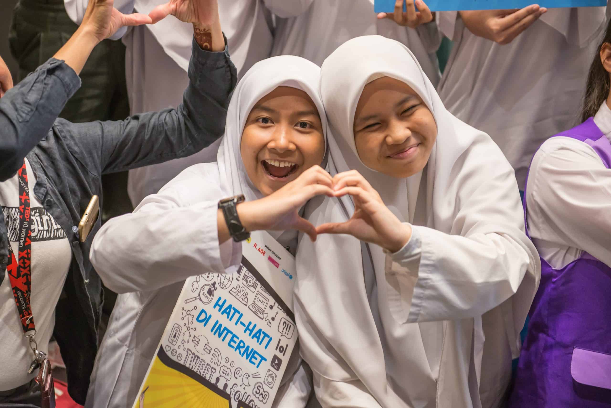 Two girls hold up their hands to make the shape of a heart. One is holding a poster that says 'hati-hati di internet'.