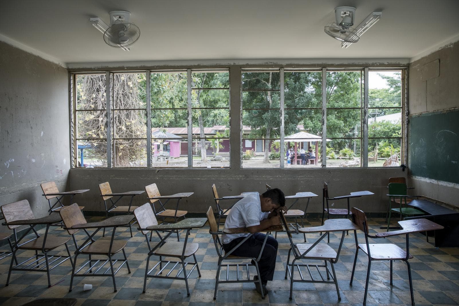 a boy who is a victim of violence sits alone in his empty classroom. He has his head on the table.