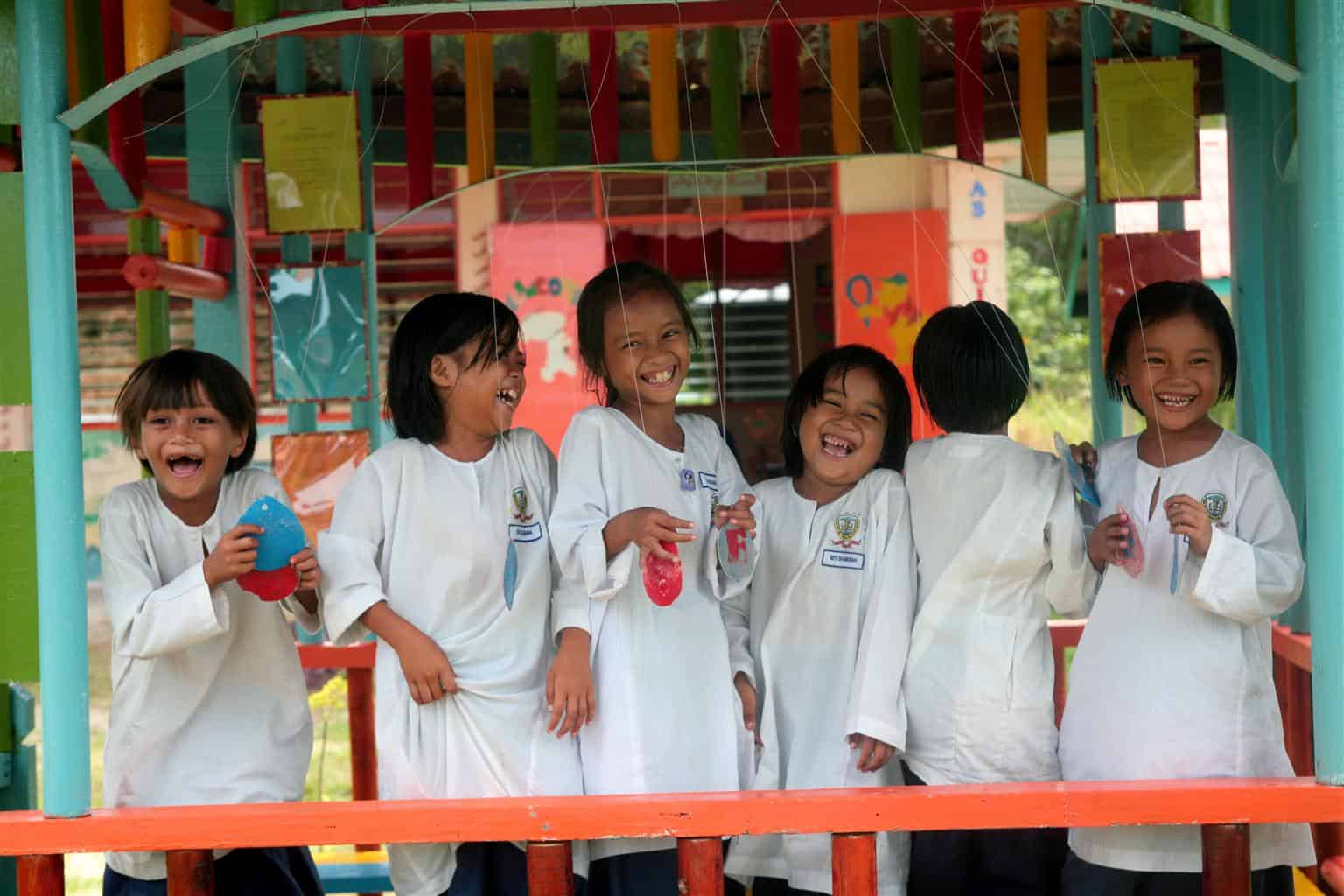 six primary school girls stand in a row outside their classroom. They are smiling and look happy.