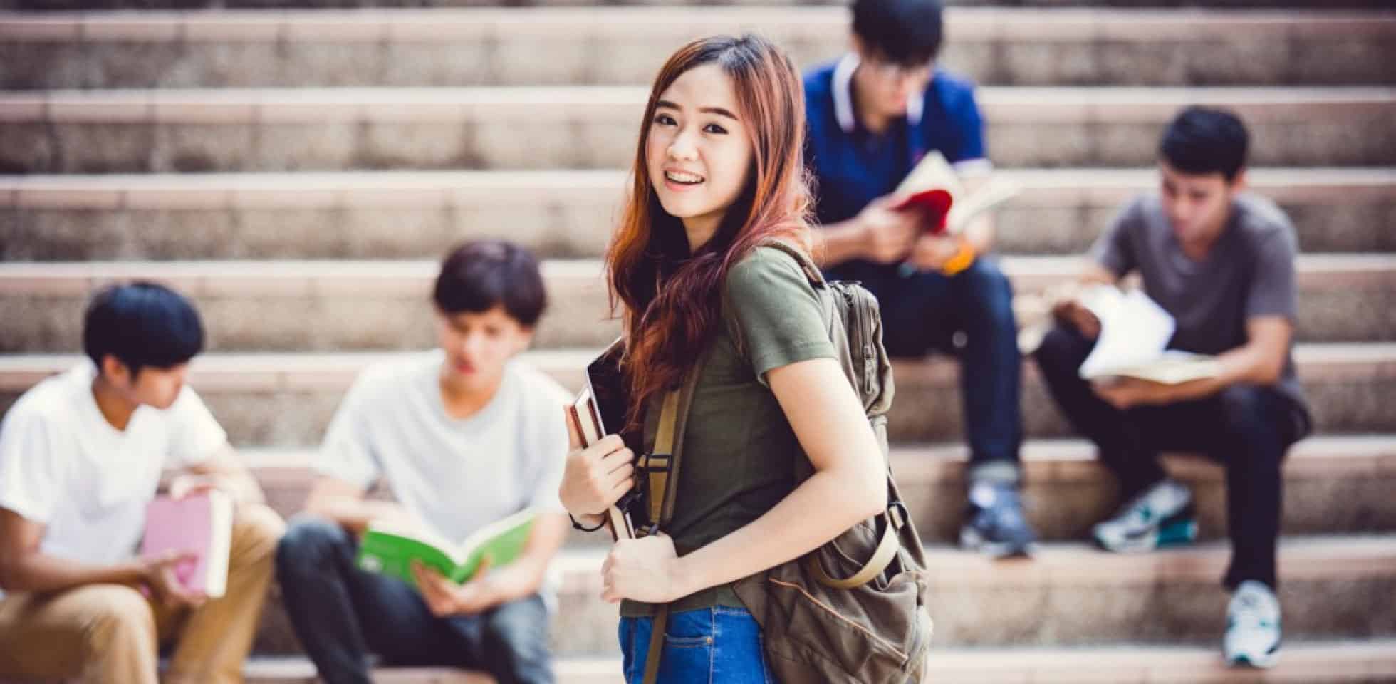 Uncovering school hacks with a college student holds her books. Behind her are some other students sitting on the stairs.