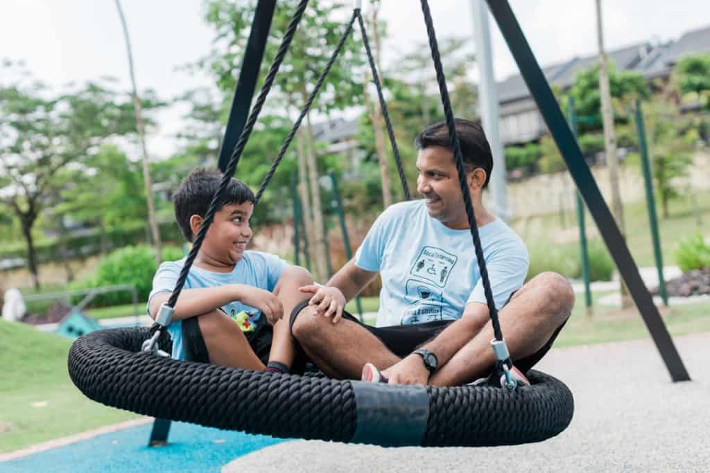 A boy and his father sitting on a special safe swing at the Inclusive Playground.