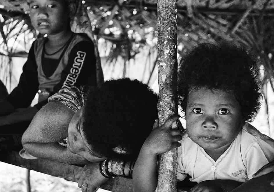 THREE YOUNG CHILDREN SITTING UNDER A HUT LOOKING AT THE CAMERA