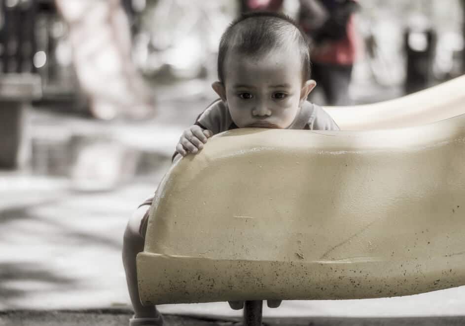 A YOUNG TODDLER BOY SITTING ON A PLAYGROUND LOOKING AT THE CAMERA