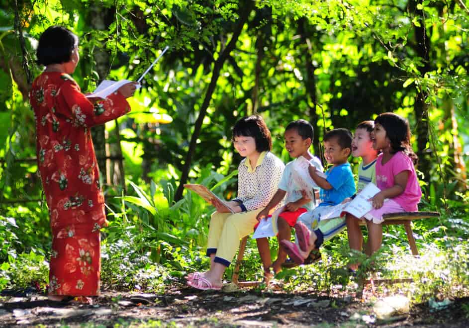 AN ADULT FEMALE TEACHER TEACHING A GROUP OF YOUNG BOYS AND GIRLS OUTSIDE