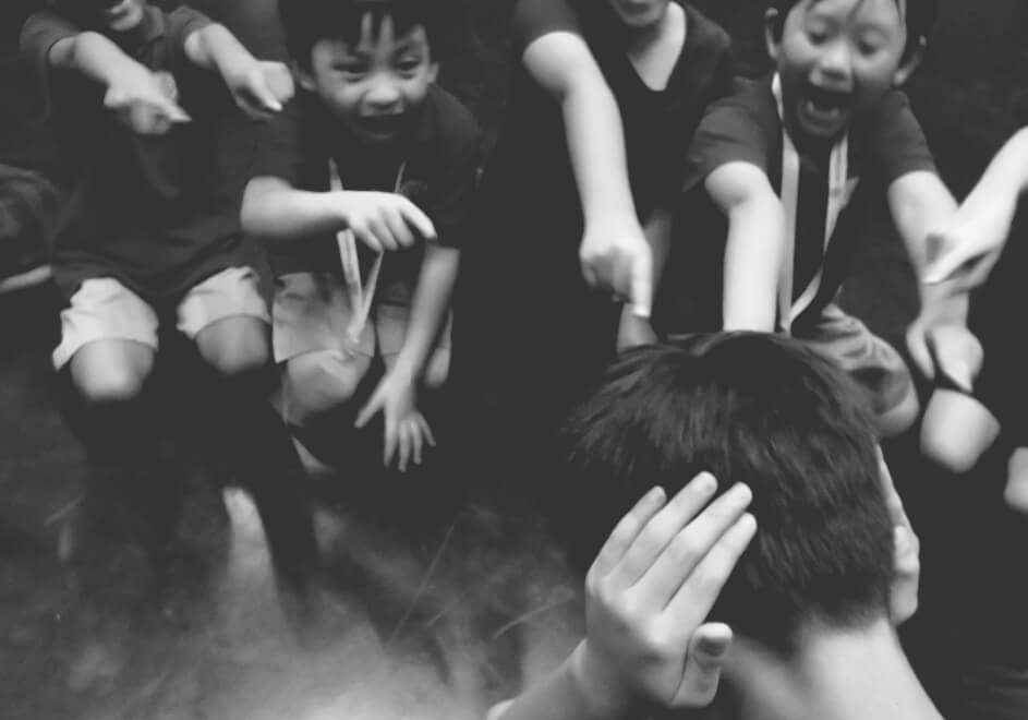 A GROUP OF YOUNG BOYS LAUGHING AND POINTING AT ANOTHER BOY WITH HIS EARS CLOSED.