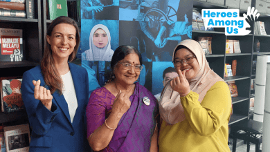 Three women stand together showing the love sign in support of the Social Work Profession Bill in Malaysia. At the corner is the Heroes Among Us logo.