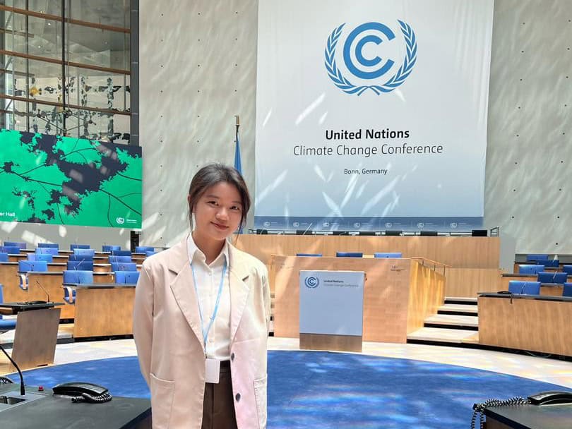 A young woman stands in front of a backdrop that has the United Nations Conference on Climate - COP - logo.