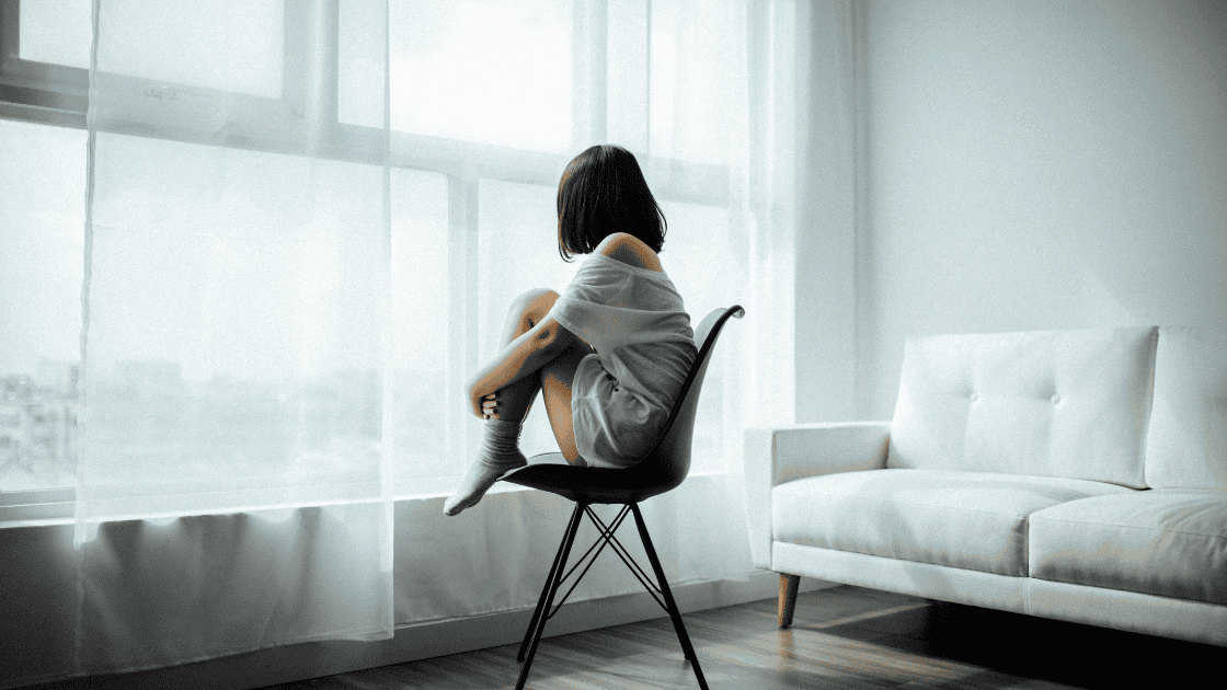 A sad young woman sitting on a chair hugging her legs.