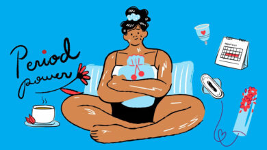 A girl sits with a hot water bottle clutched to her tummy. She is surround with sanitary products around her.