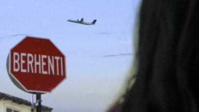 A person looks up at a plane flying in the sky. A signboard says "Berhenti" (Stop).