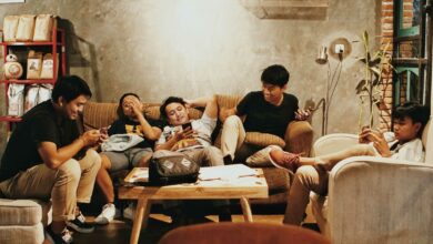 Five young people relax in a shared indoor space, each looking at a mobile phone while seated around a coffee table. The image represents the role of group chats and democracy in shaping conversations, sharing information and civic engagement among young people.