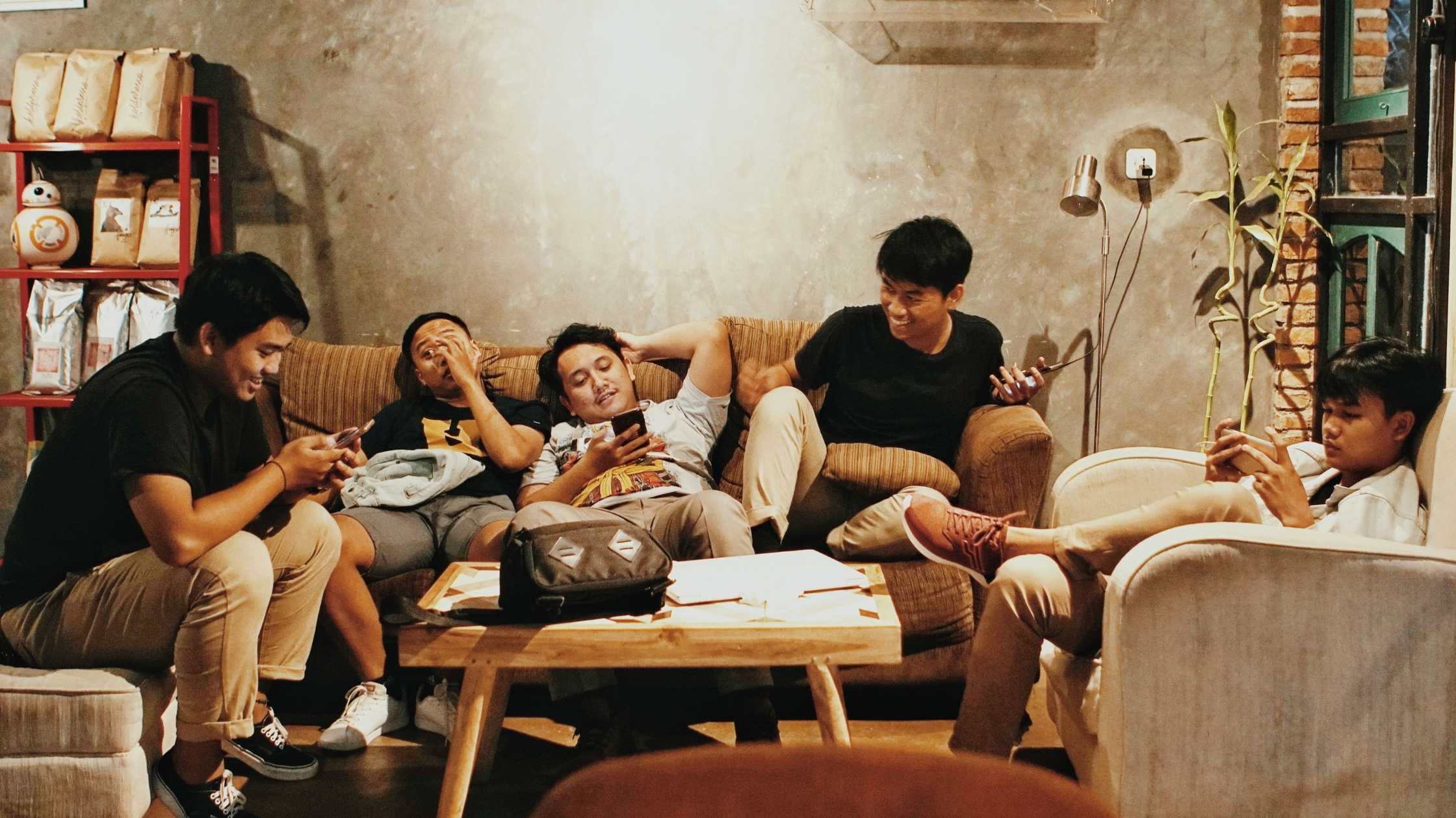 Five young people relax in a shared indoor space, each looking at a mobile phone while seated around a coffee table. The image represents the role of group chats and democracy in shaping conversations, sharing information and civic engagement among young people.